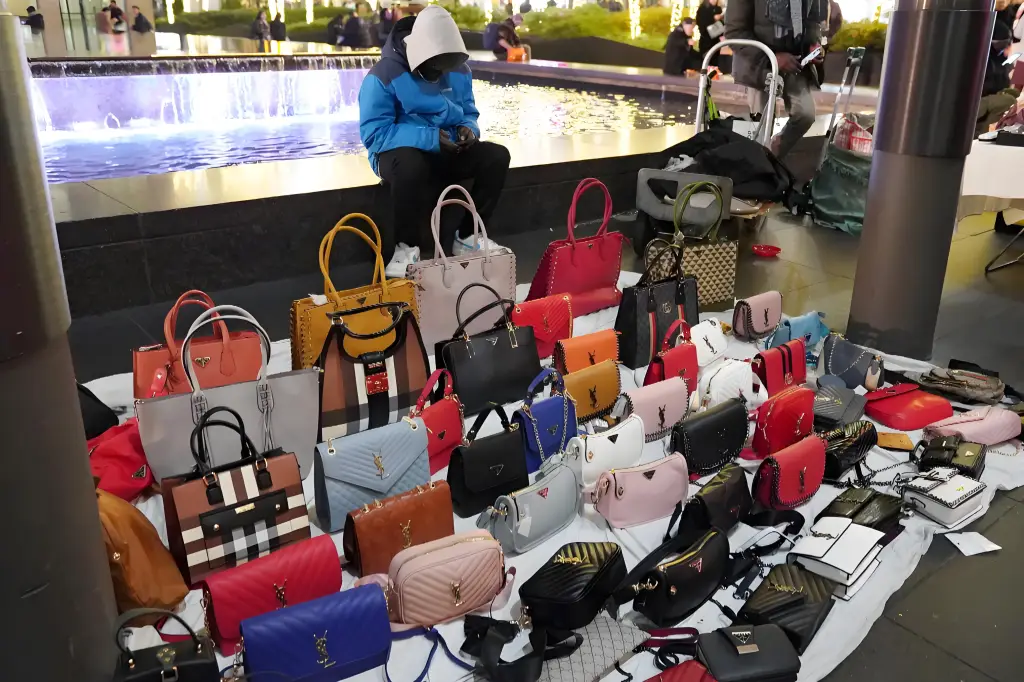 A street vendor with a large assortment of colorful designer - style handbags laid out for sale near a fountain in an urban setting, likely featuring replica bags.