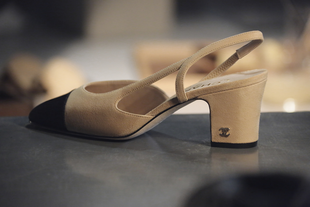 A side view of a beige suede and black leather two-tone slingback shoe with a small Chanel interlocking CC logo on the heel, representing iconic Chanel designs.